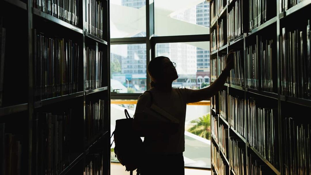 Female student in silhouette looking at books on a bookshelf
