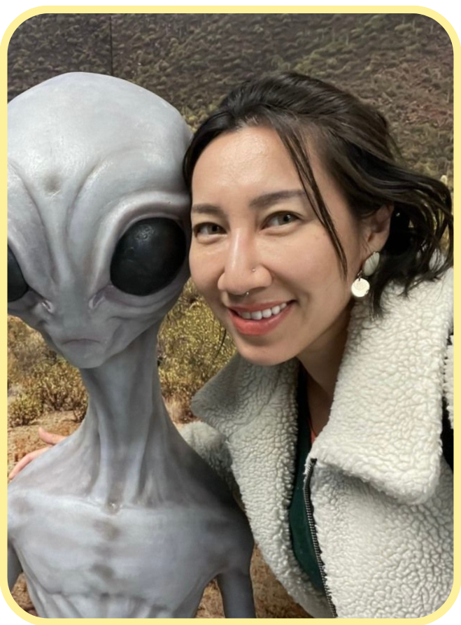 Portrait photograph of Dr. Shana Ye is seen smiling beside a mannequin resembling a science fiction alien character. 