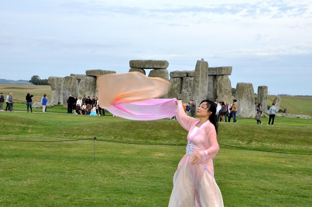 A person in a pink dress with a scarf in front of a stone monument

Description automatically generated