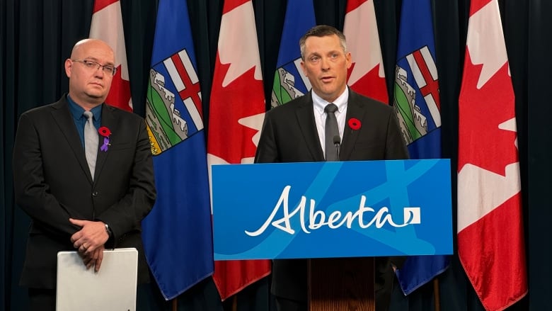 Finance Minister Nate Horner, wearing a black suit, white shirt, and dark grey tie, stands at a lectern with the blue Alberta logo on it, in front of a series of Canadian and Alberta flags. Minister Searle Turton stands on Horner's right-hand side, wearing a black suit, dark blue shirt and silver tie. Both men are wearing poppies on their lapels and Turton is holding a folder.